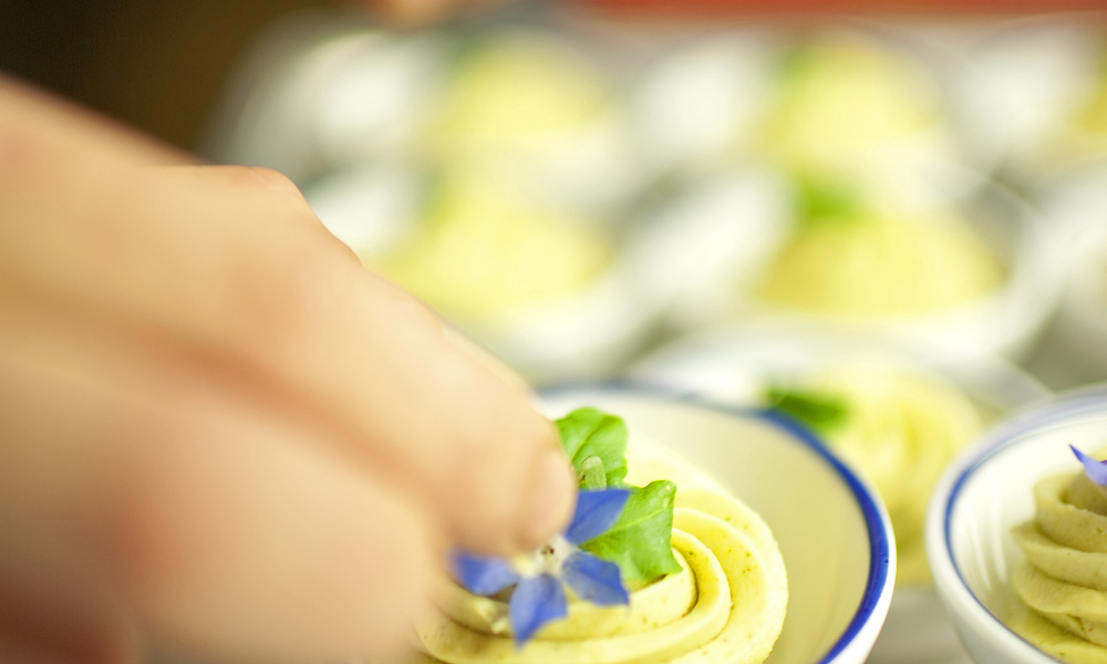Eine Hand fügt eine violette essbare Blume zu einer gelben Mousse in einer kleinen Schale hinzu.