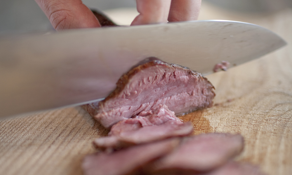 Eine Hand schneidet mit einem großen Messer ein medium-rare Steak auf ein Holzbrett.