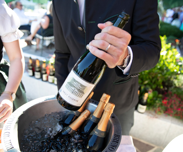 A person in a suit holds a bottle of sparkling wine over an ice bucket containing other bottles.