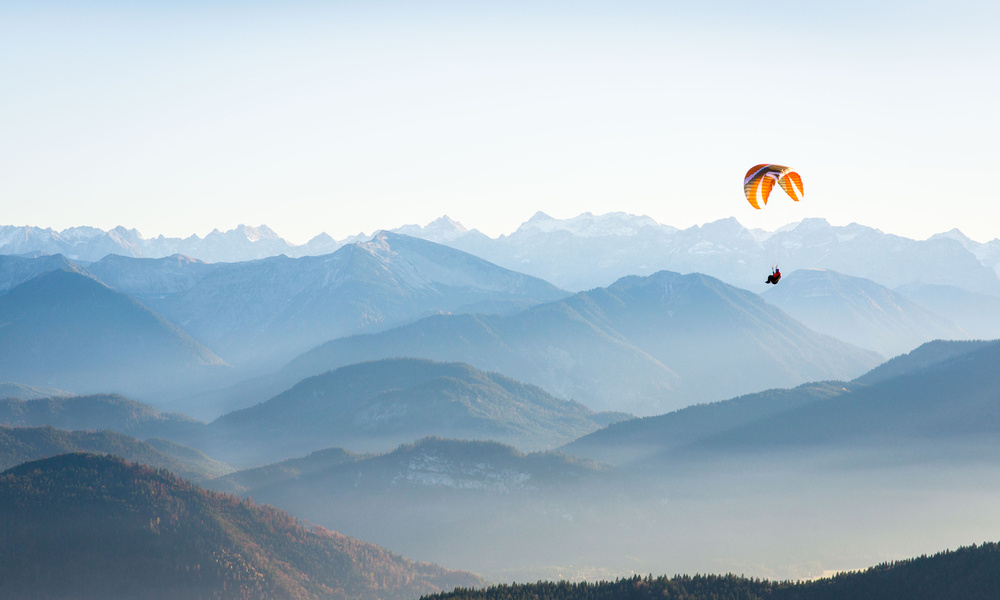 Ein Gleitschirmflieger schwebt über nebligen blauen Bergen unter einem klaren Himmel.