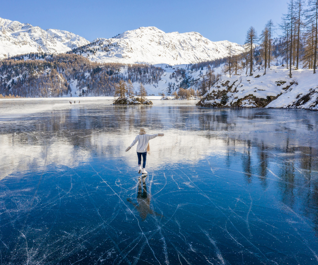 Eine Person läuft Schlittschuh auf einem zugefrorenen See, umgeben von schneebedeckten Bergen und Bäumen unter einem klaren blauen Himmel.
