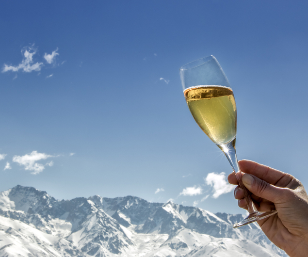 Eine Hand hält ein Glas Champagner mit verschneiten Bergen und einem klaren blauen Himmel im Hintergrund.