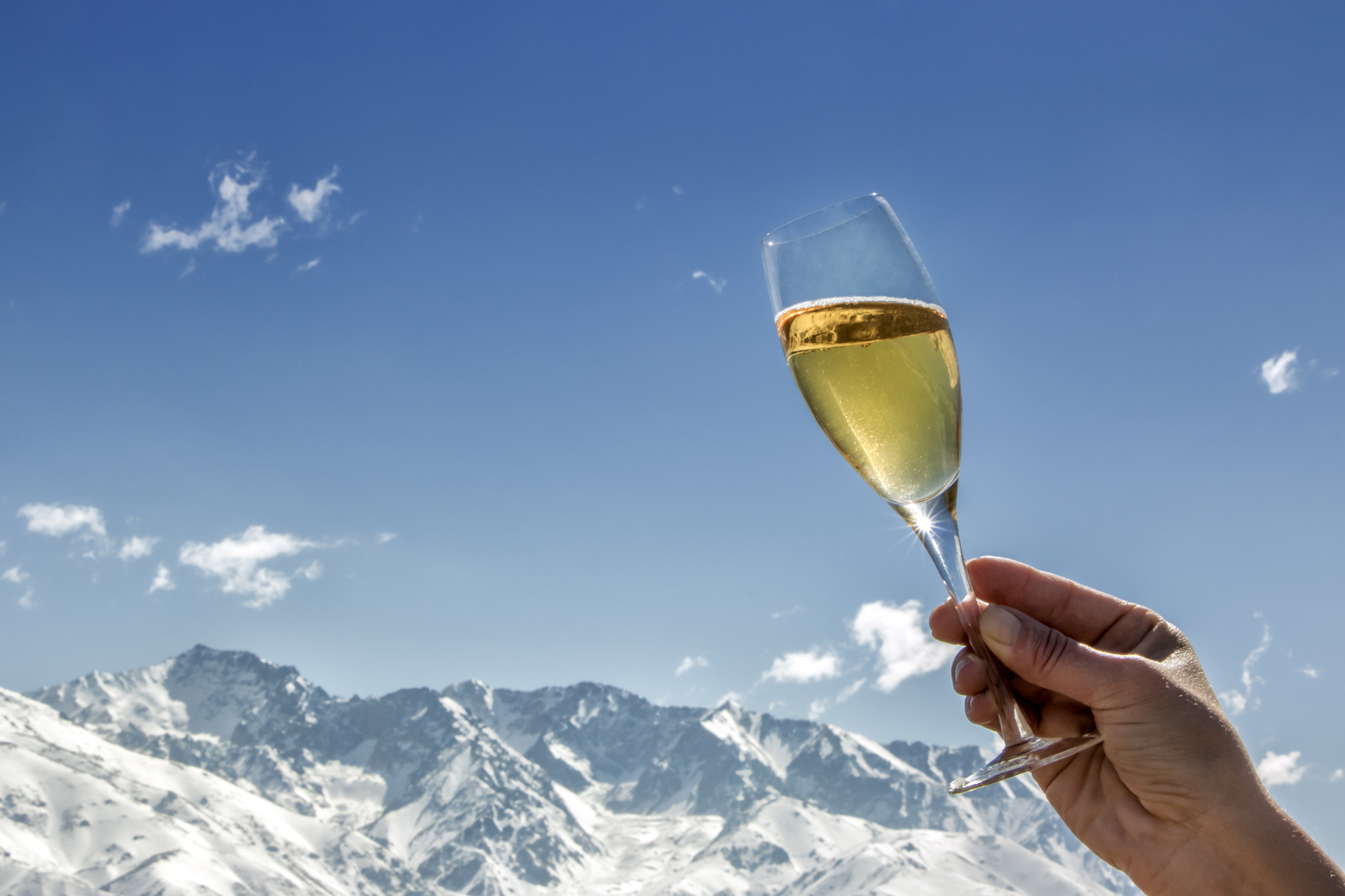 One hand holds a glass of champagne with snow-covered mountains and a clear blue sky in the background.