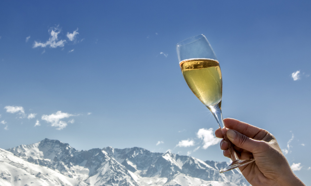 Eine Hand hält ein Glas Champagner mit verschneiten Bergen und einem klaren blauen Himmel im Hintergrund.