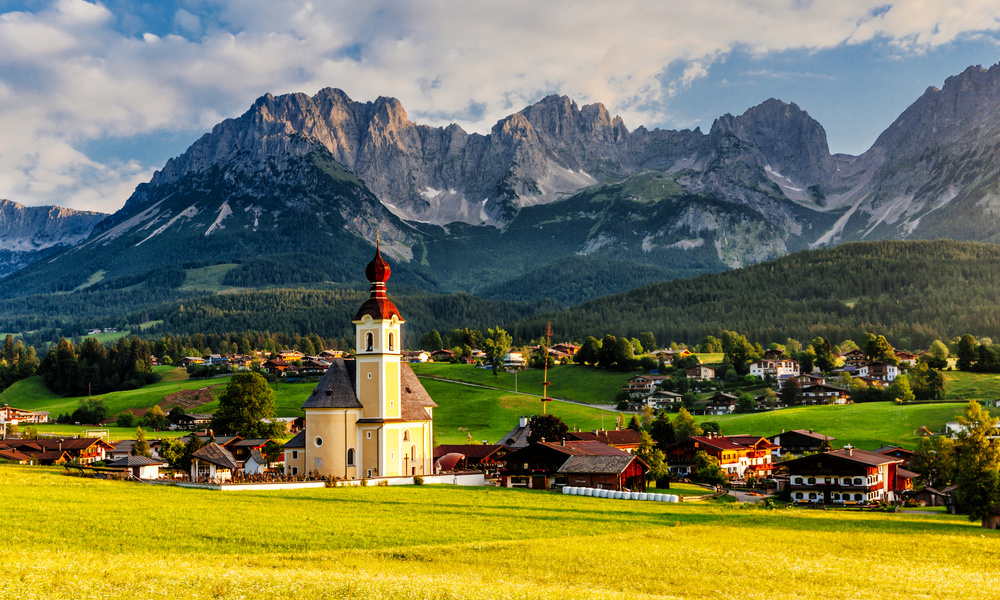 Eine Kirche und ein Dorf mit alpinen Bergen im Hintergrund unter einem teilweise bewölkten Himmel.