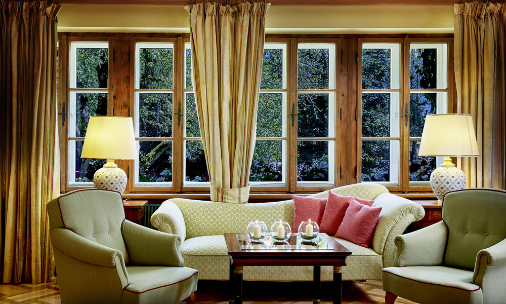 Elegantes Wohnzimmer mit Sesseln, Sofa, Lampen und großen Fenstern mit Vorhängen, die die Bäume draußen zeigen.