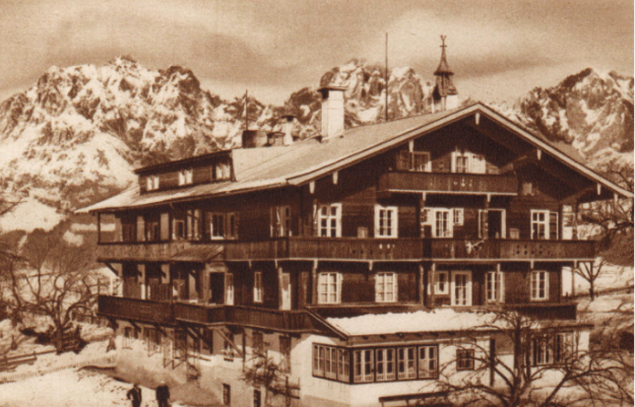 A large wooden chalet stands in front of snow-covered mountains with bare trees and snow on the ground.
