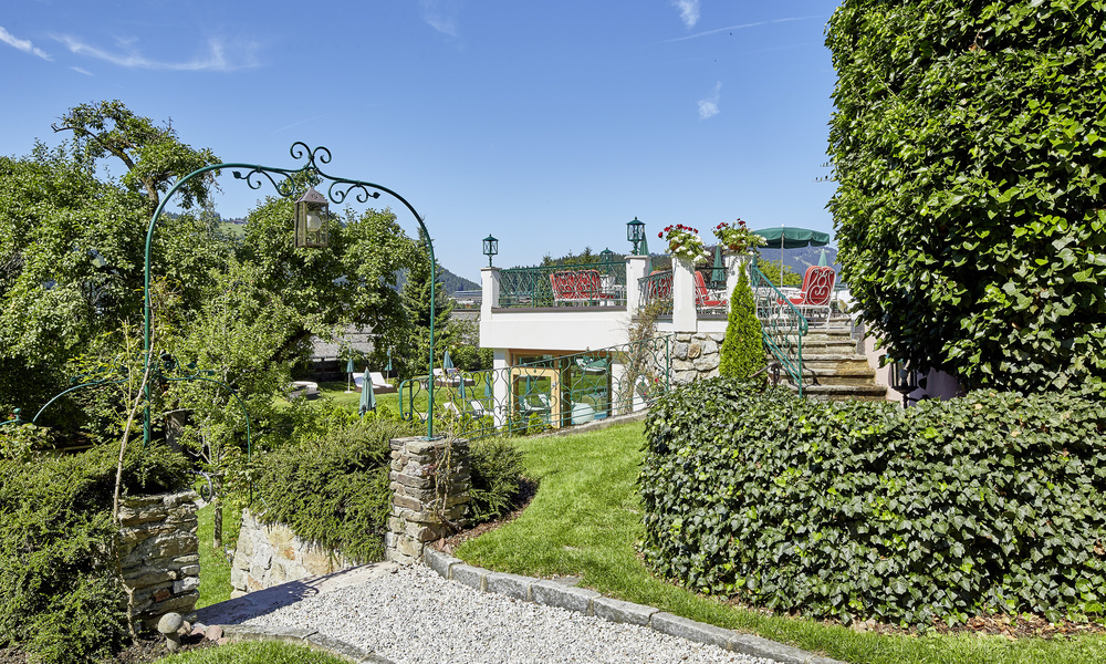 Gartenweg mit Steinstufen, schmiedeeisernem Bogen, grünen Hecken und einer Terrasse mit roten Stühlen unter blauem Himmel.