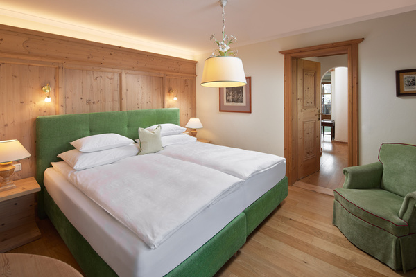Gemütliches Hotelzimmer mit grünem Bett, Holzvertäfelung, Sessel, Lampen und offenem Durchgang zu einem anderen Zimmer.