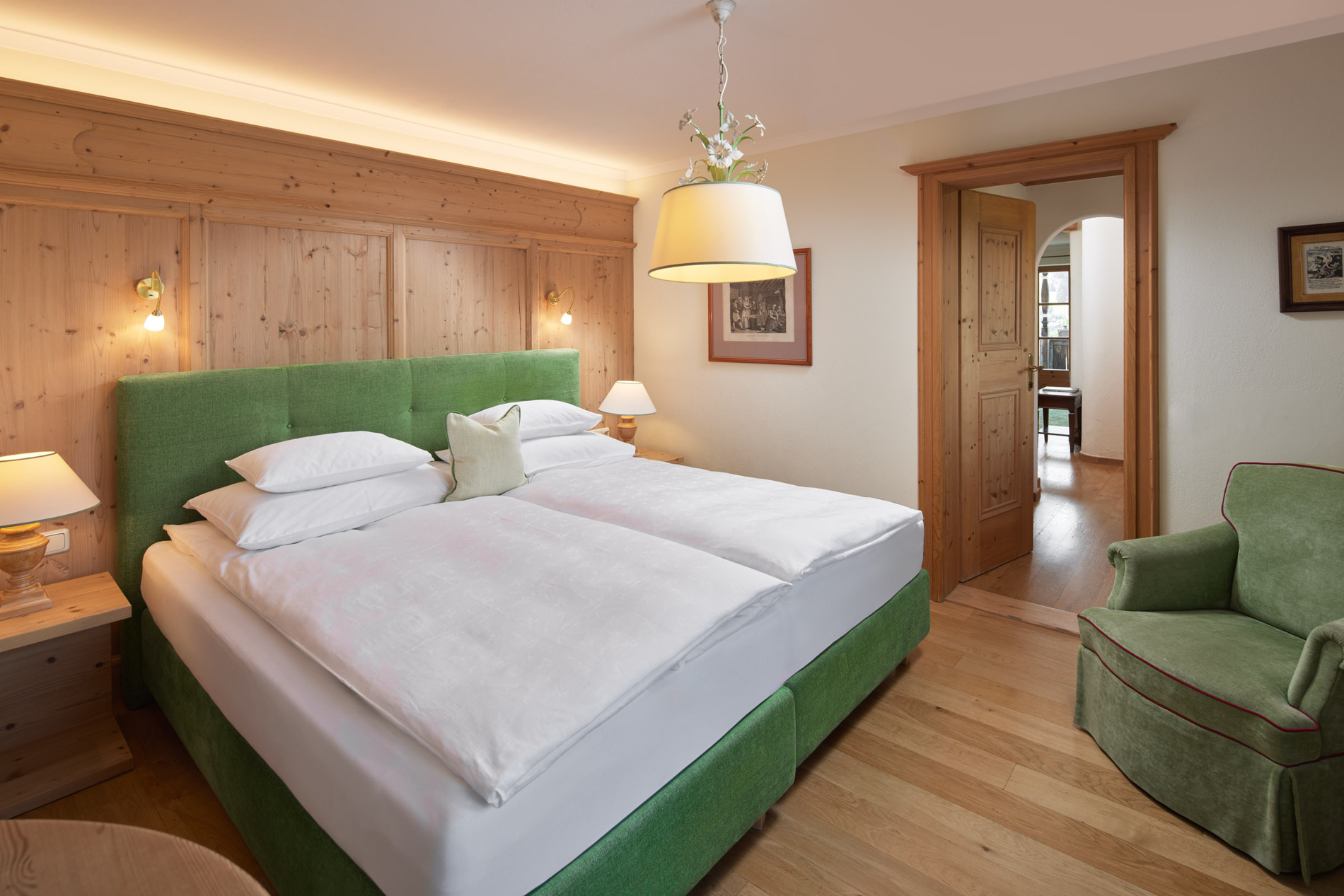 Gemütliches Hotelzimmer mit grünem Bett, Holzvertäfelung, Sessel, Lampen und offenem Durchgang zu einem anderen Zimmer.