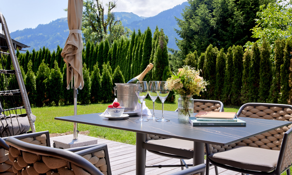 Terrassentisch im Freien mit Champagner, Gläsern, Blumen und Büchern vor einer malerischen Kulisse aus Bergen und Grünpflanzen.
