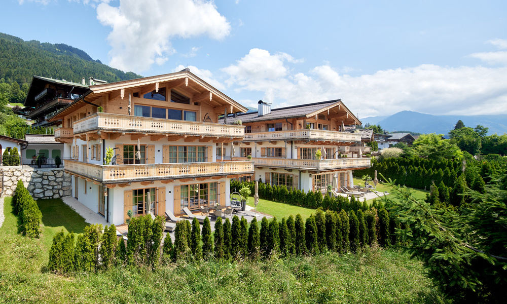 Zwei große Holzchalets mit Balkonen, umgeben von Grün und Bergen unter blauem Himmel.