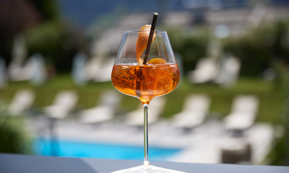 Ein Glas Orangencocktail mit Eis und einer Orangenspirale, im Freien an einem Swimmingpool.