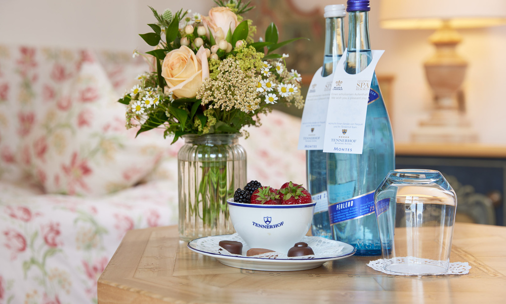 Eine Vase mit Blumen, Wasser in Flaschen, eine Schale mit Beeren und Pralinen auf einem Holztisch in einem gemütlichen Raum.