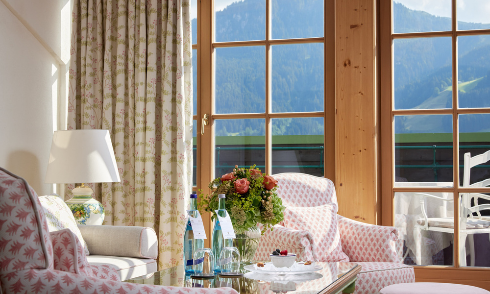 Helle Sitzecke mit geblümten Sesseln, einer Lampe und Blumen, mit Blick auf die Berge durch große Fenster.