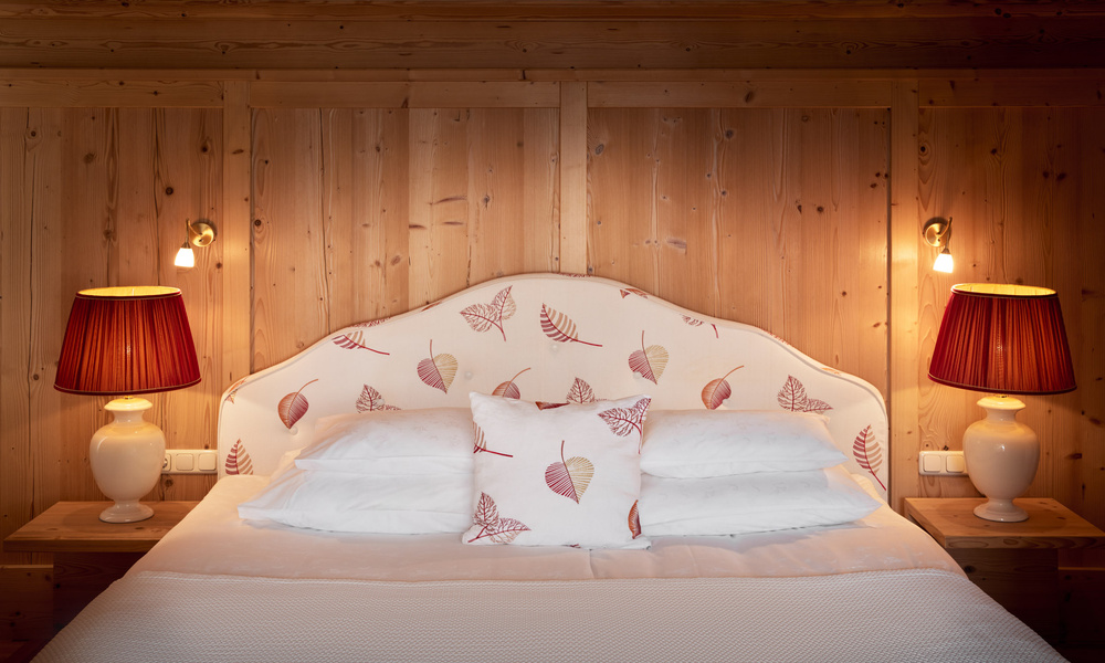Gemütliches Bett mit blattgemusterten Kissen, flankiert von roten Lampen und Holzverkleidung an der Wand dahinter.