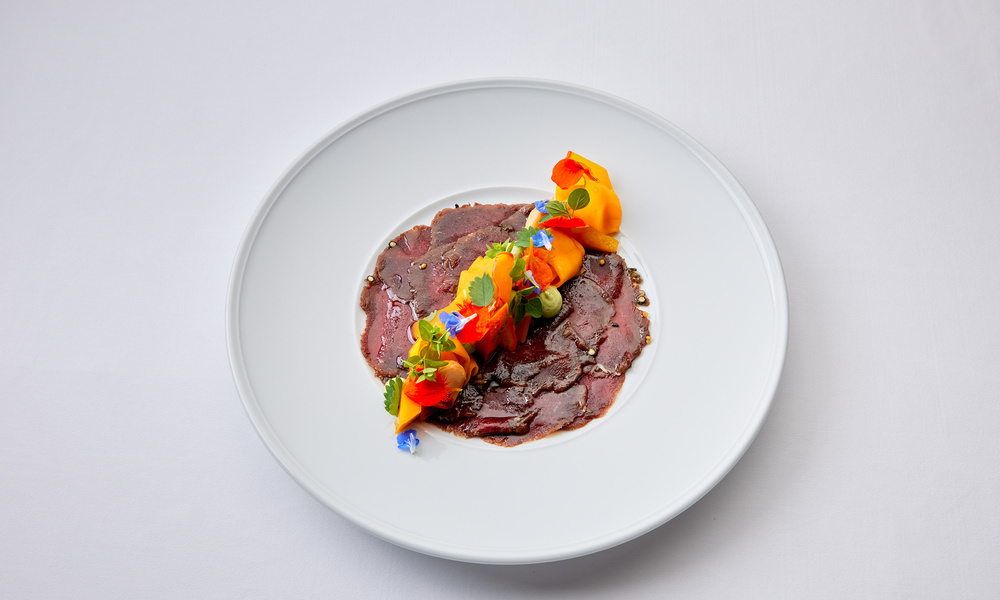 Dünne Fleischscheiben, garniert mit bunten essbaren Blumen und Kräutern auf einem weißen Teller.