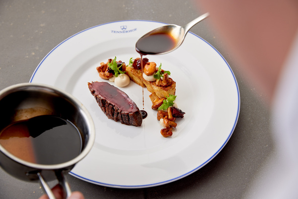 Ein Koch gießt Sauce über ein Gourmet-Steakgericht mit Beilagen auf einem weißen Teller.