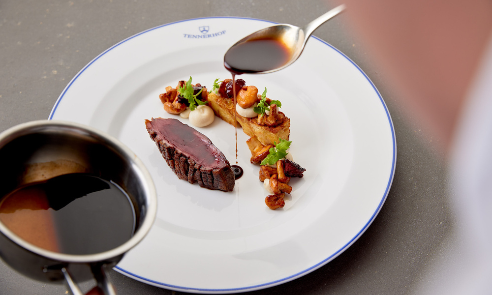 Ein Koch gießt Sauce über ein Gourmet-Steakgericht mit Beilagen auf einem weißen Teller.