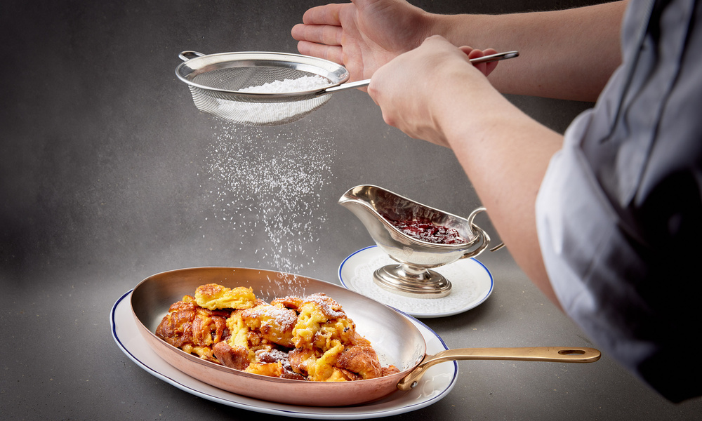 Eine Person streut Puderzucker über eine Dessertschale mit einer Sauciere im Hintergrund.