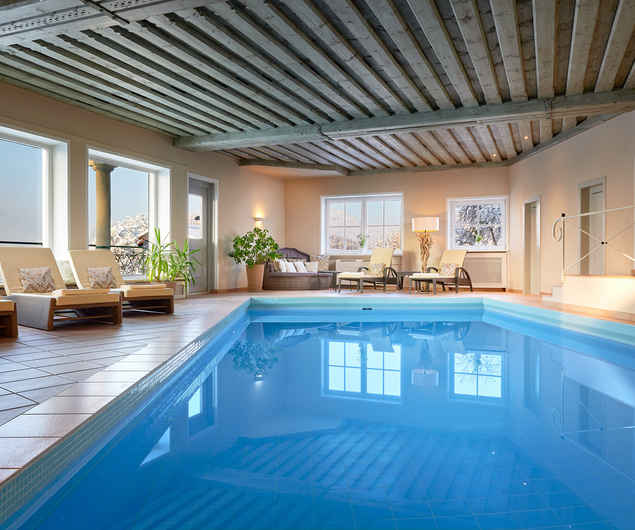 Indoor pool with sun loungers, potted plants and large windows reflecting the clear water.