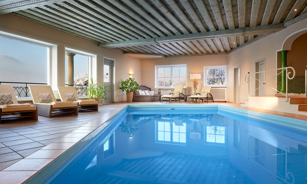 Innenpool mit Liegestühlen, Topfpflanzen und großen Fenstern, in denen sich das klare Wasser spiegelt.