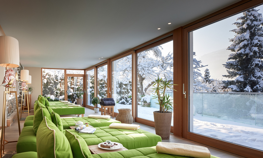 Moderne Spa-Lounge mit grünen Sesseln an großen Fenstern mit Blick auf eine verschneite Winterlandschaft mit Kiefern.