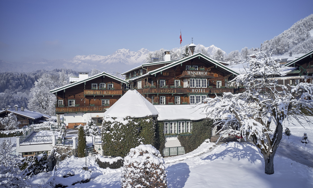 Ein verschneites Alpenhotel aus Holz, umgeben von schneebedeckten Bäumen und Bergen unter einem strahlend blauen Himmel.