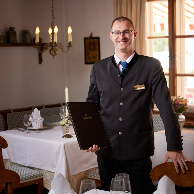 A smiling waiter in a suit with a menu in his hand stands in a neatly furnished, elegant restaurant dining room.