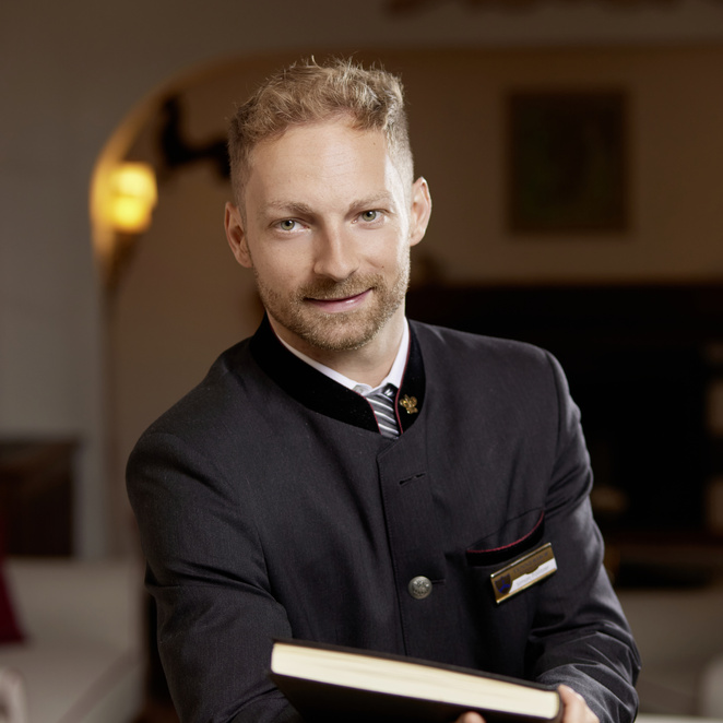 Smiling man in a dark uniform jacket holding a book in a warmly lit interior.