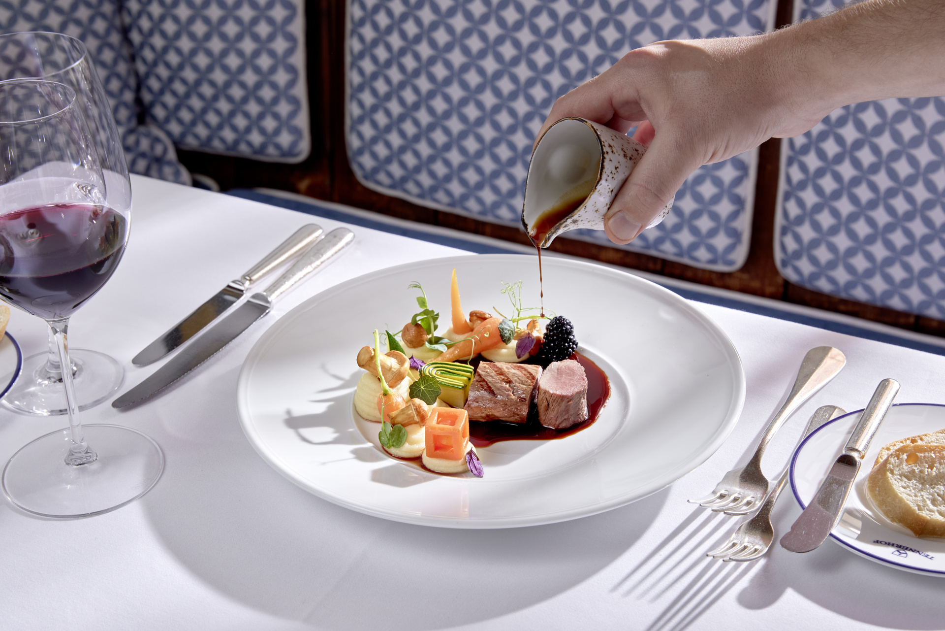 Eine Hand gießt Soße über ein Gourmet-Fleischgericht, dazu Wein und Brot auf einem weißen Tischtuch.