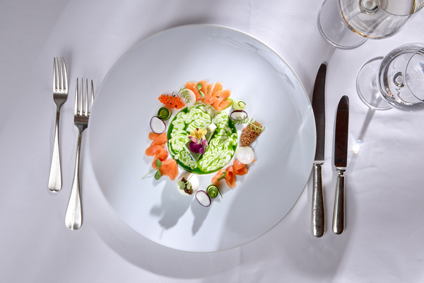 Ein Feinschmeckersalat, kunstvoll angerichtet auf einem weißen Teller mit Besteck und Gläsern auf einem weißen Tischtuch.