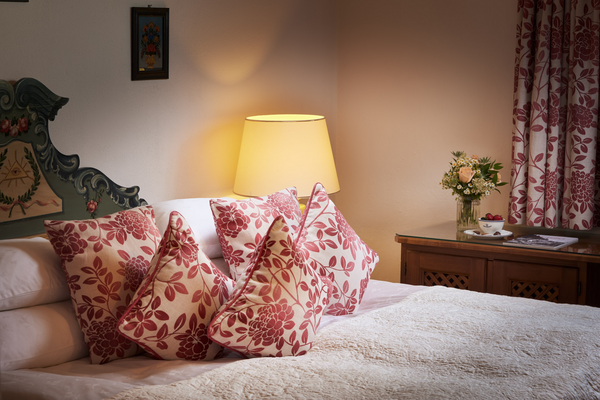 Cozy bedroom with floral pillows, a lighted lamp and a bedside table with flowers and a teacup.