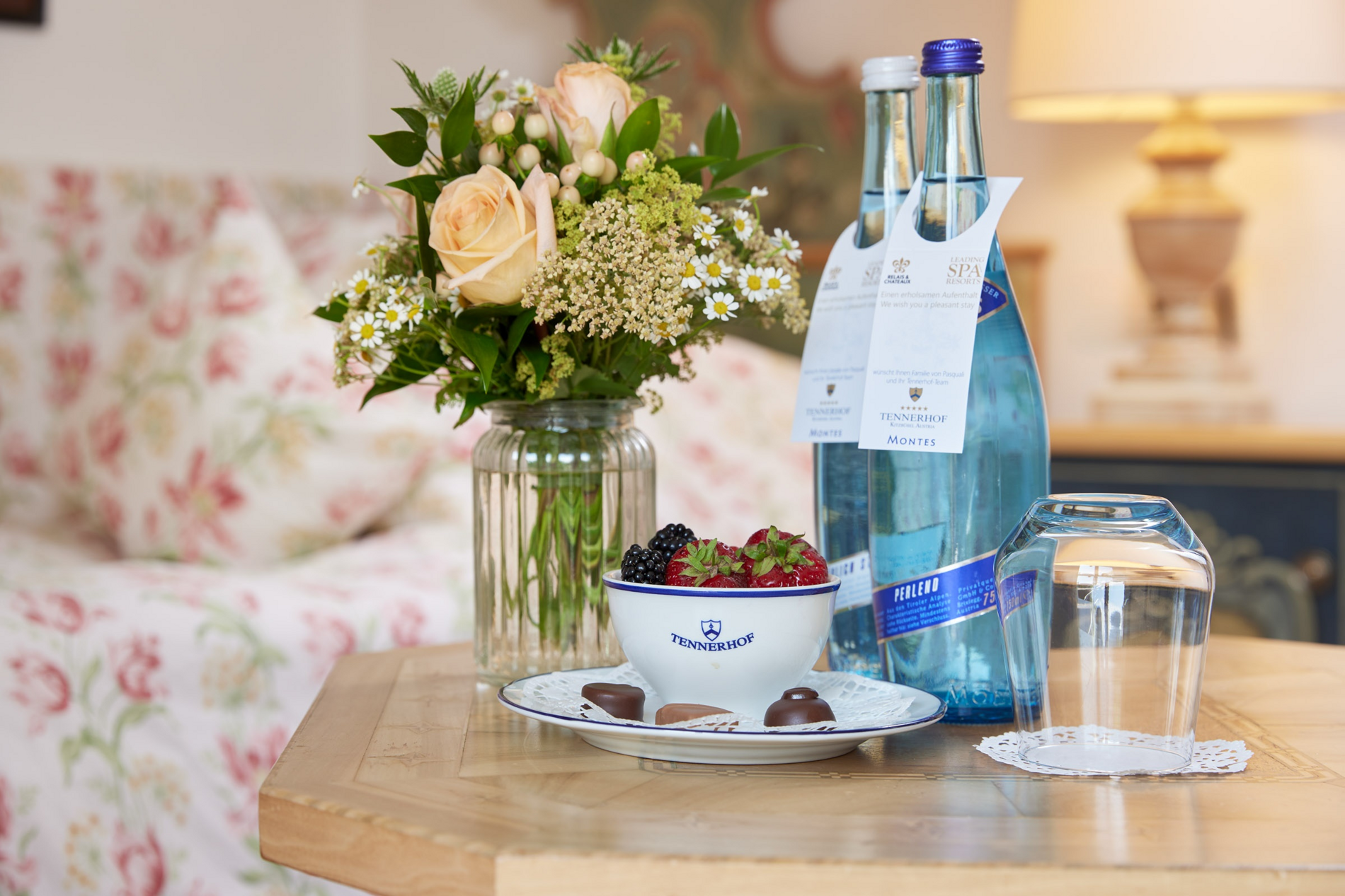 Eine Schale mit Beeren, Schokolade, Wasser in Flaschen und ein Glas auf einem Tisch mit einer Blumenvase im Hintergrund.