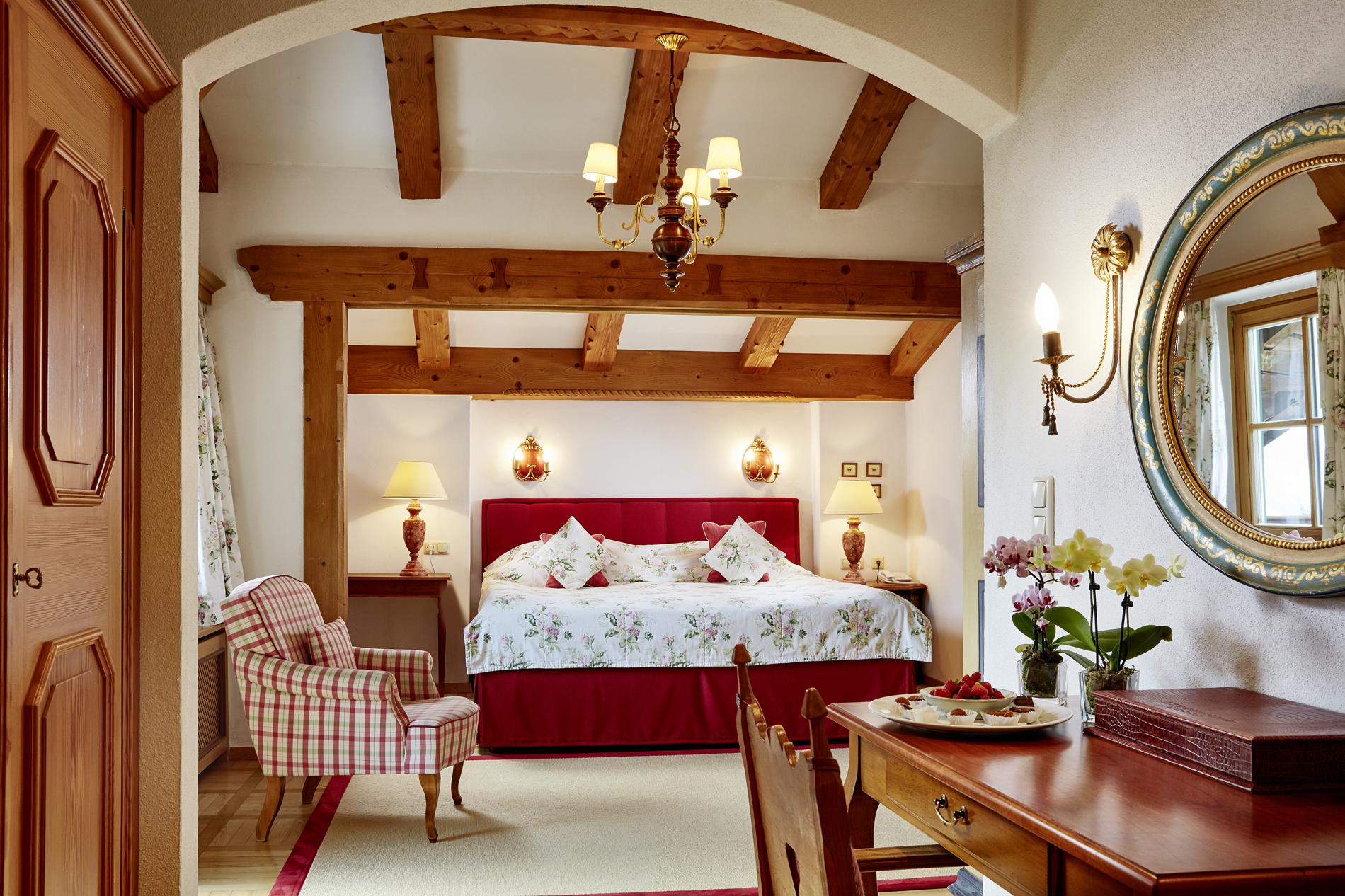 Cozy bedroom with a flowered bed, wooden beams, a striped chair and a round mirror above a desk.