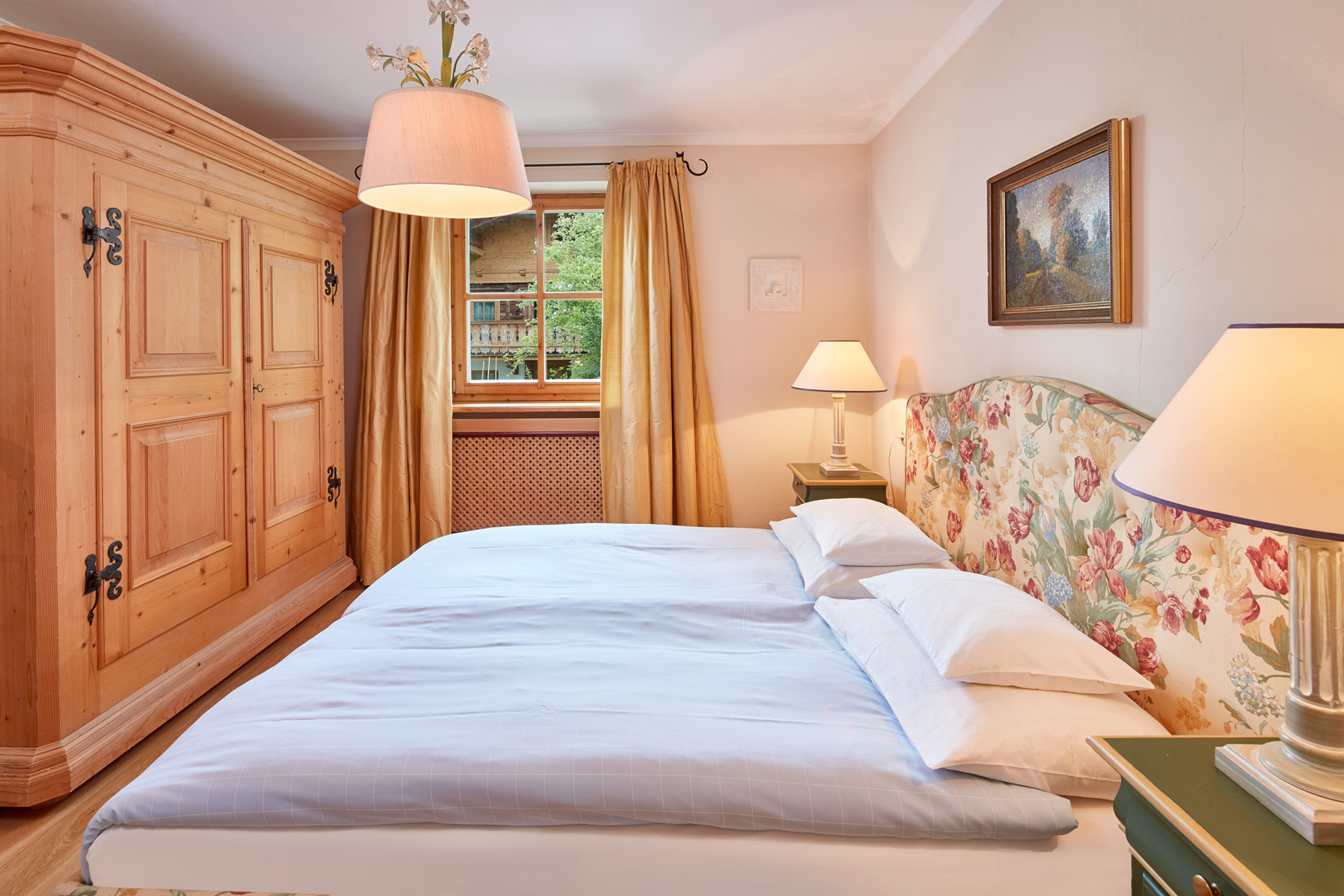 Gemütliches Schlafzimmer mit geblümtem Kopfteil, Doppelbett, Kleiderschrank aus Holz und warmer Beleuchtung durch Lampen.