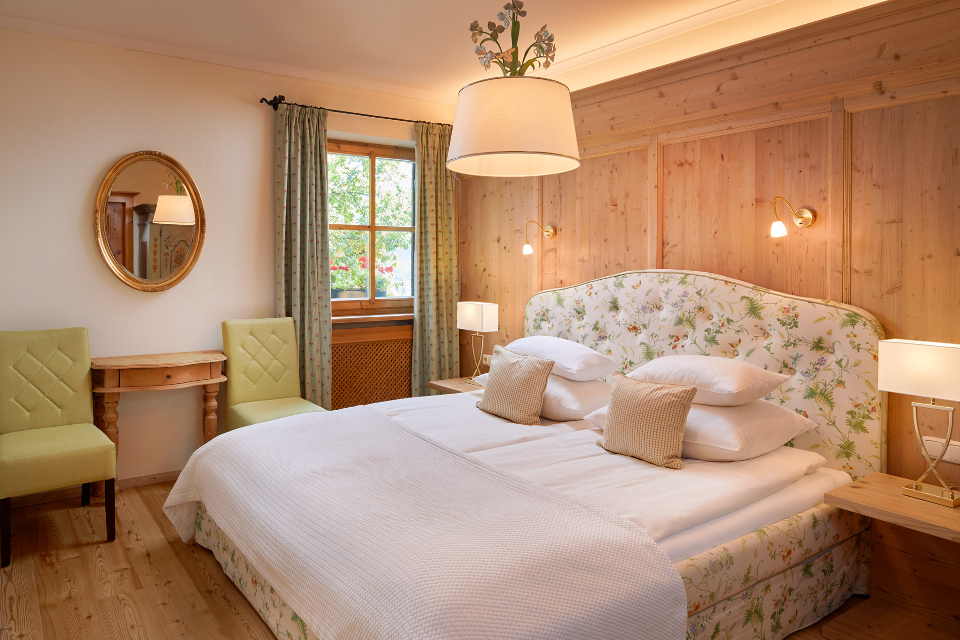Cozy bedroom with floral decor, a double bed, two green chairs, wooden walls and a window with curtains.